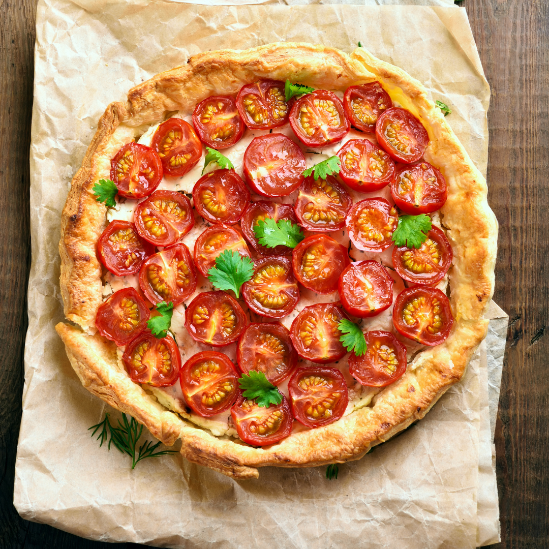 A round tomato tart with a golden-brown crust, topped with sliced red tomatoes and small green herb garnishes, presented on parchment paper over a dark wooden surface.