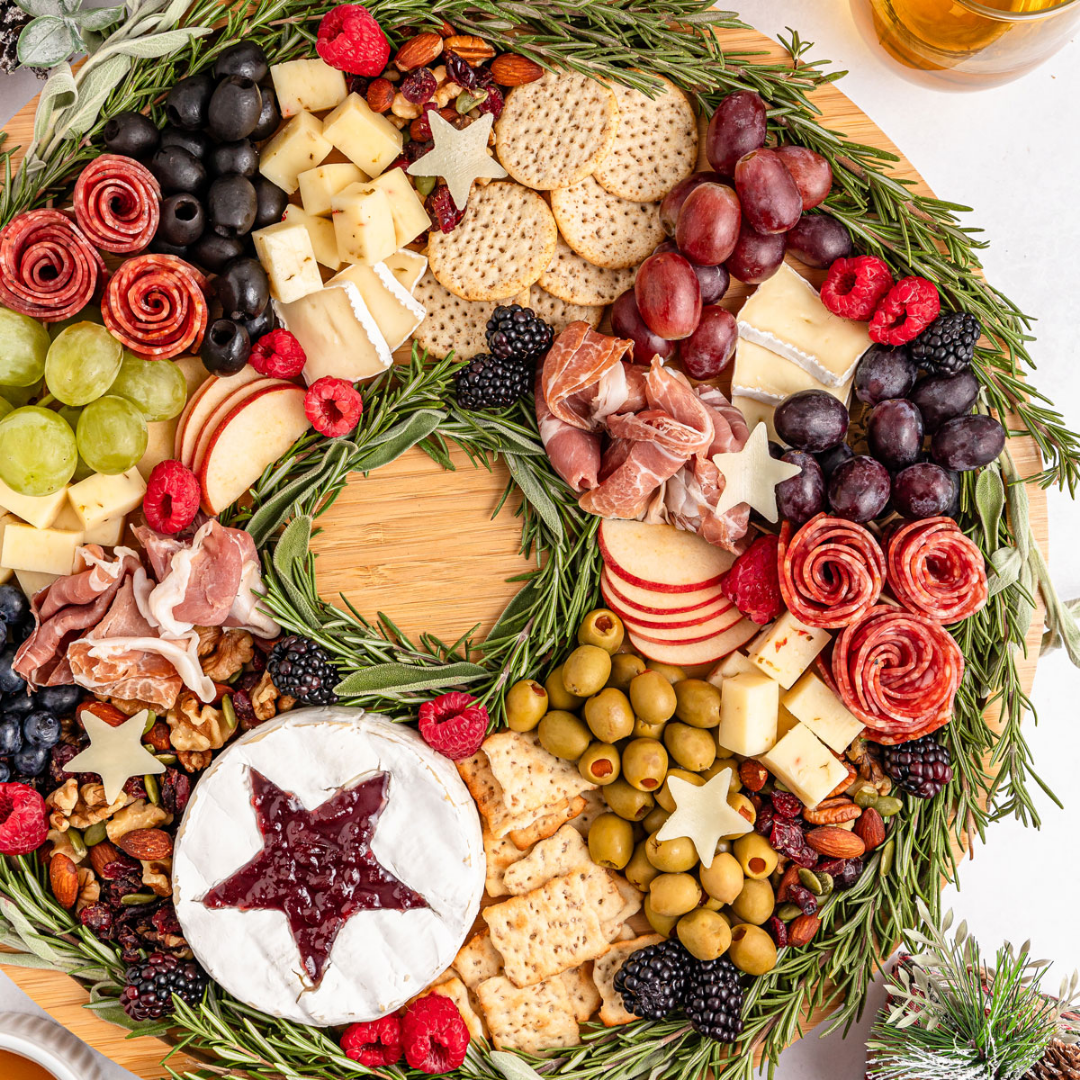 Holiday wreath charcuterie board ringed with rosemary, with brie topped with a jam star, salami roses, prosciutto, olives and berries