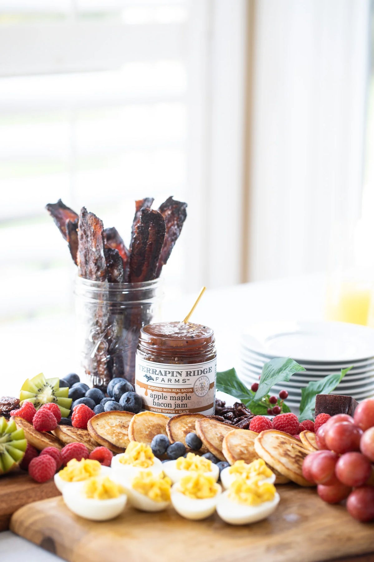 Assorted food items including crackers, fruits, and a jar of jam on a wooden board with Apple Maple Bacon Jam