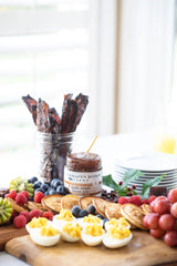 Assorted food items including crackers, fruits, and a jar of jam on a wooden board with Apple Maple Bacon Jam