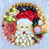 Santa-face charcuterie board with a cheese face, brie hat and mustache, salami rose, prosciutto, fresh fruit and olives