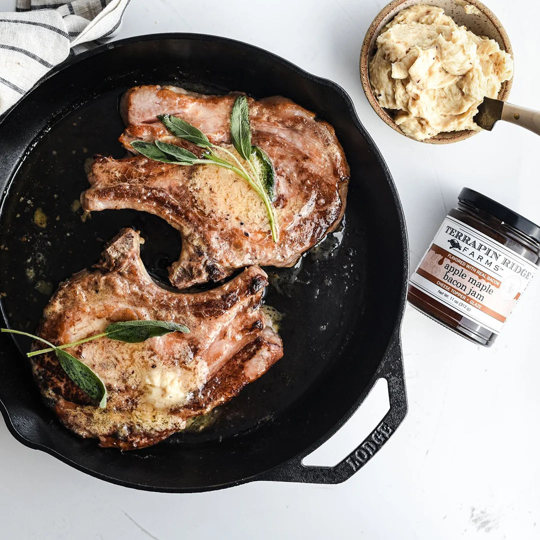 Searing pork chops with herbs in a cast iron skillet on a white surface, with a jar of apple maple bacon jam nearby.