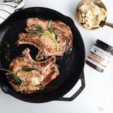 Searing pork chops with herbs in a cast iron skillet on a white surface, with a jar of apple maple bacon jam nearby.